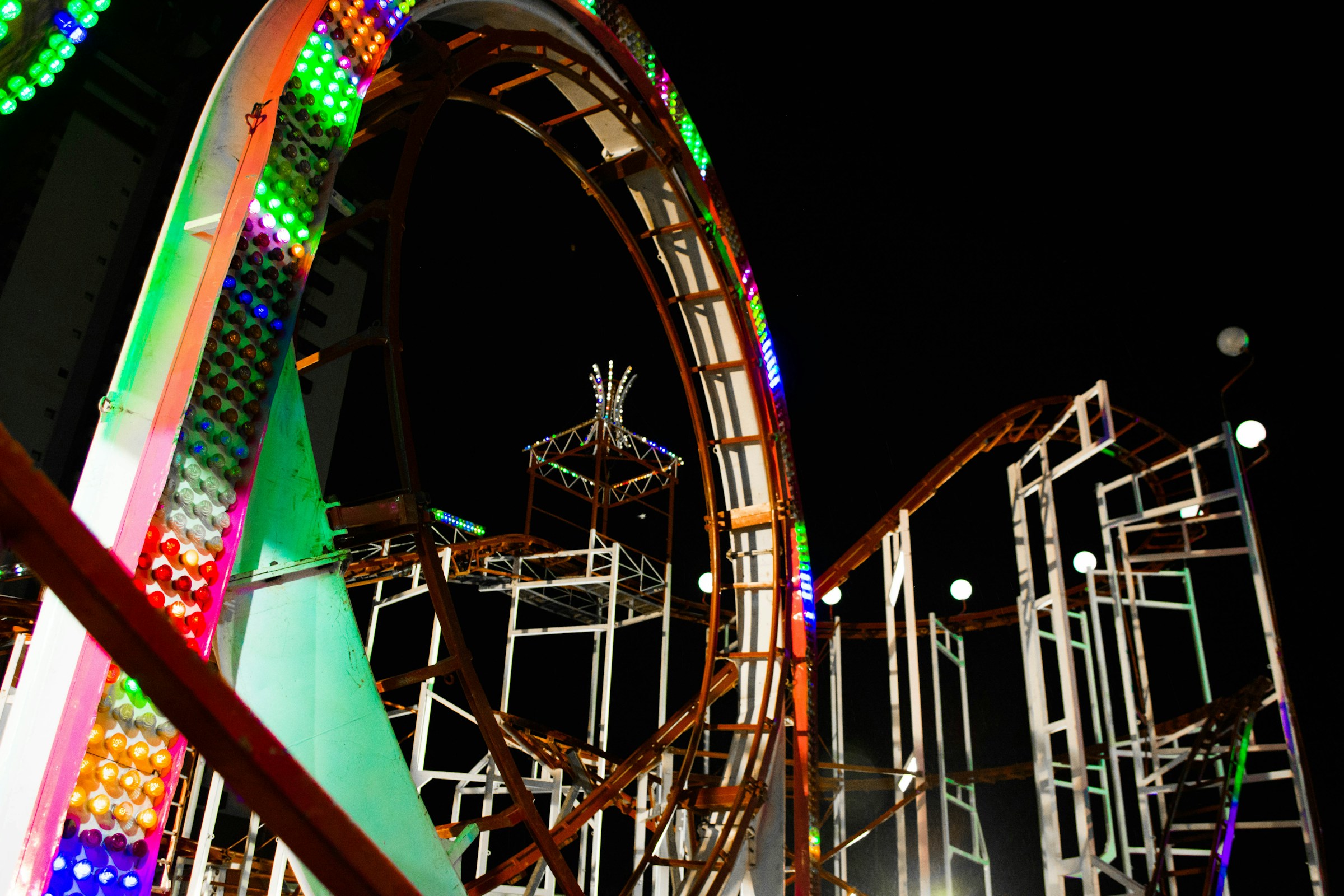A looping fairground roller coaster photographed from below at night, its circular loop and framework outlined in rows of bright green, pink, orange, and blue bulbs, with white-lit track snaking away to the right against a pitch-black sky.